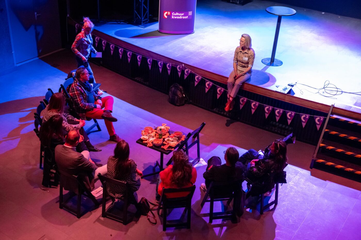 intieme theatersetting, spreker zittend op podium, kleine kring van 10 toehoorders